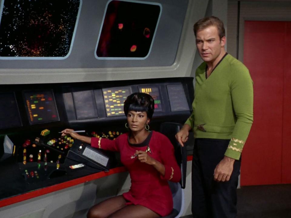 Nichelle in a red dress uniform and William in a green shirt universe. Both are at a control panel.