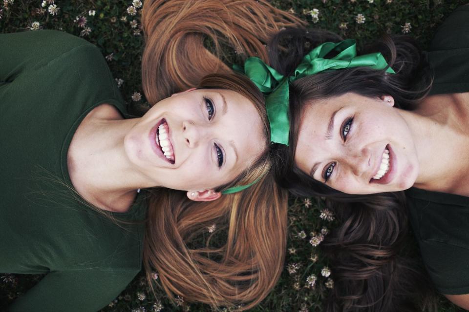 two girls laying down on the grass laughing upwards towards the sky it might be st patricks day because of the green bows in their hair