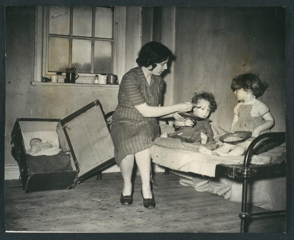 A woman sits on a bed feeding a young child, while another child stands beside them. An infant sleeps in a suitcase crib nearby