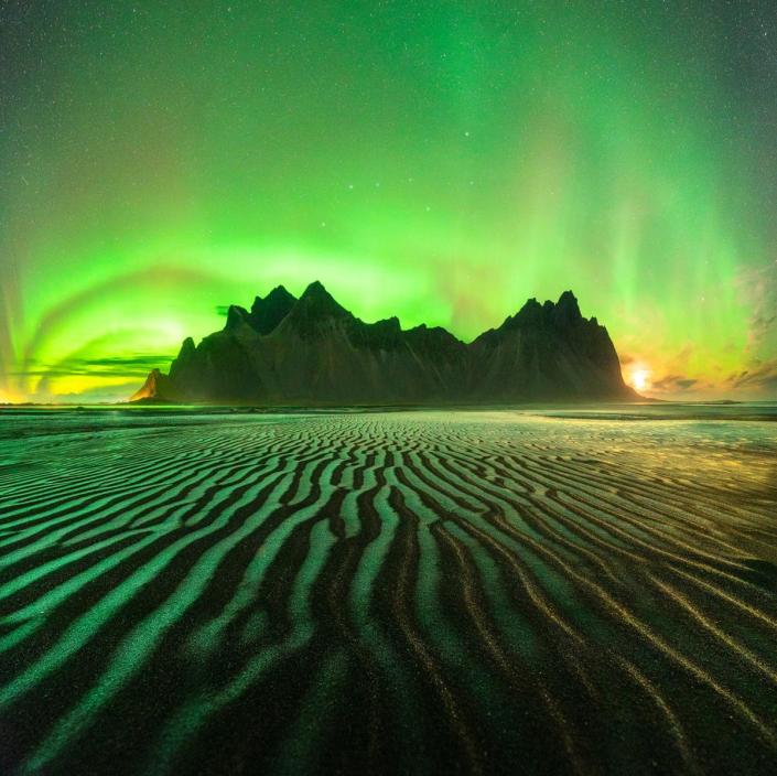 A mountain rises from a beach&#39;s wavy black sands, glowing in greens and pale yellows beneath the blazing aurora arched in the sky.