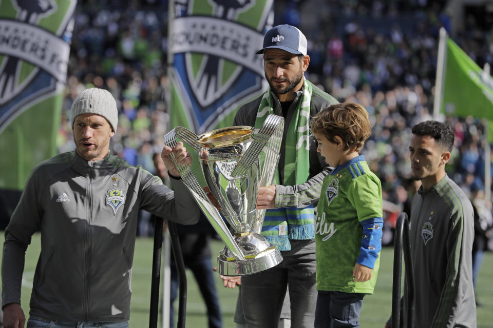 Seattle Sounders' defender Gustav Svensson, left, and midfielder Nicolas Lodeiro, center, bring in the Sounders' 2019 MLS Cup trophy with the help of Lodeiro's son Leandro, second from right, before the start of their season-opening an MLS soccer match against the Chicago Fire, Sunday, March 1, 2020, in Seattle. (AP Photo/Ted S. Warren)