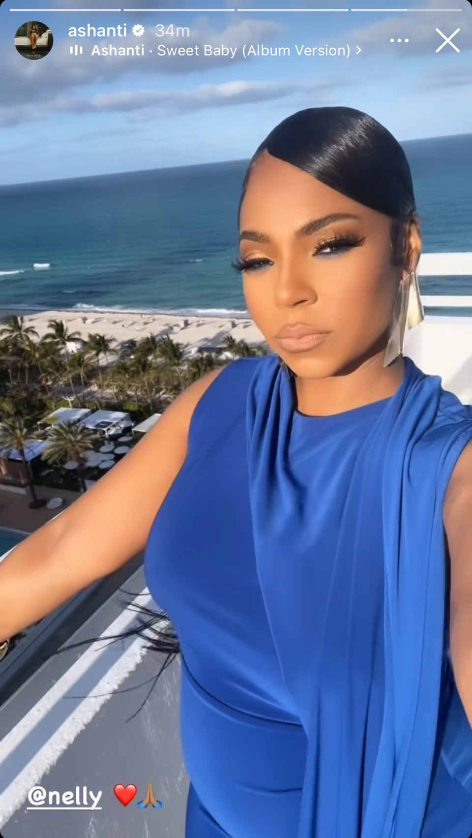 Ashanti poses in a sleeveless blue gown with large earrings, palm trees, and buildings in the background