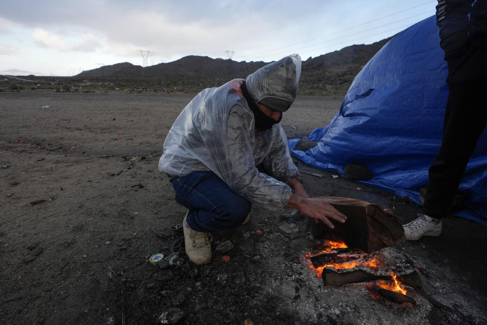An asylum-seeking migrant warms his hands as he waits to be processed in a makeshift, mountainous campsite after crossing the border with Mexico, Friday, Feb. 2, 2024, near Jacumba Hot Springs, Calif. (AP Photo/Gregory Bull)