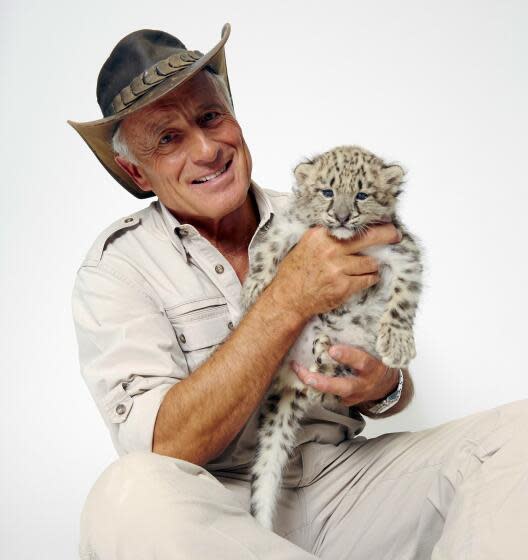 Jack Hanna wears a wide-brimmed hat and hold a snow leopard cub while sitting on the floor