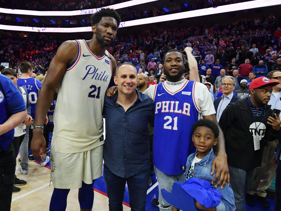 joel embiid, a tall man in a philadephia 76ers jersey, michael rubin, a shorter man in a blue untucked shirt, meek mill, another man in a philadelphia jersey, and his son papi, a young child, stand on a basketball court