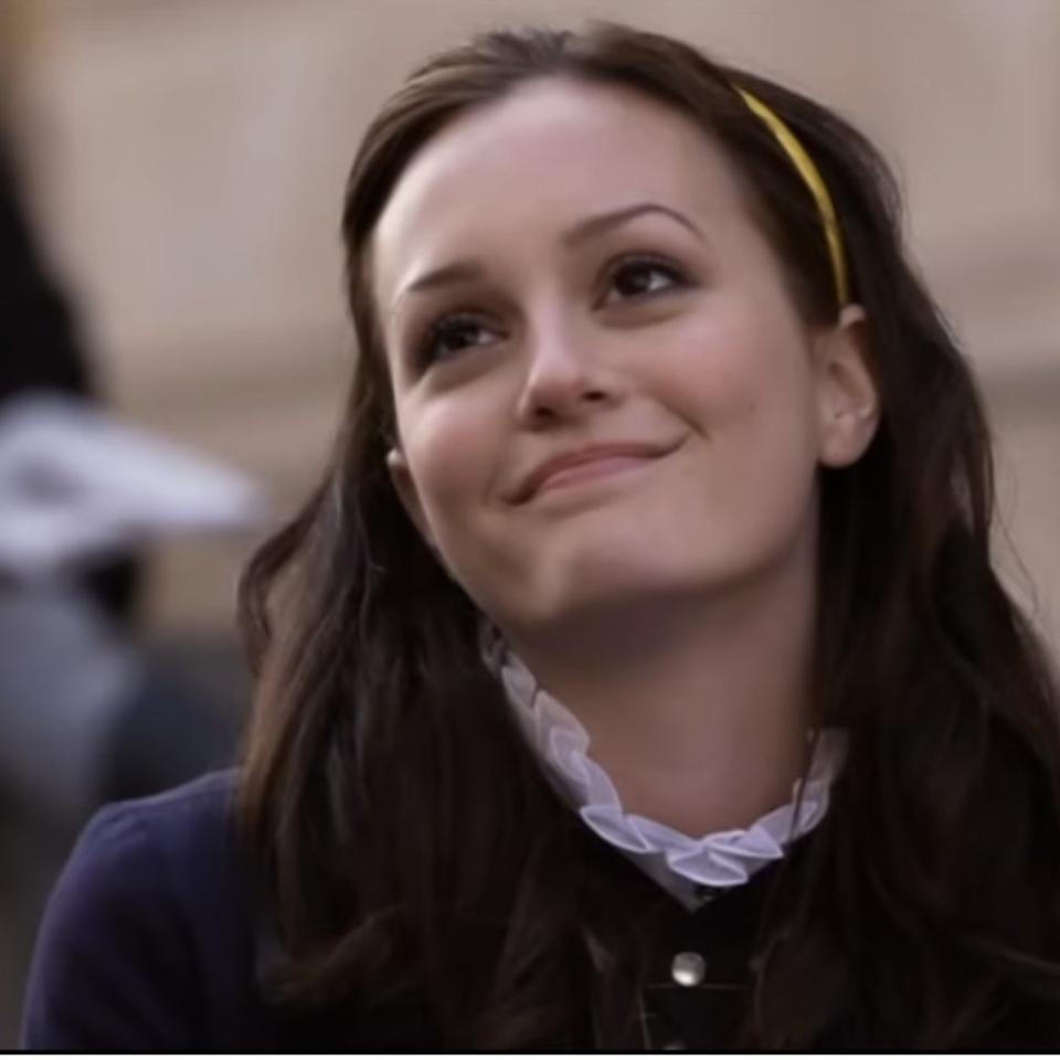 Leighton Meester is sitting and smiling while looking slightly to the side. She is wearing a headband and a blouse with a ruffled collar