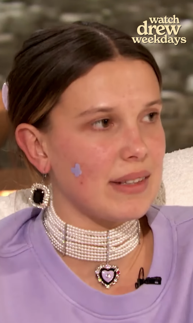 Millie Bobby Brown in a purple top with decorative earrings and a choker necklace, on the talk show