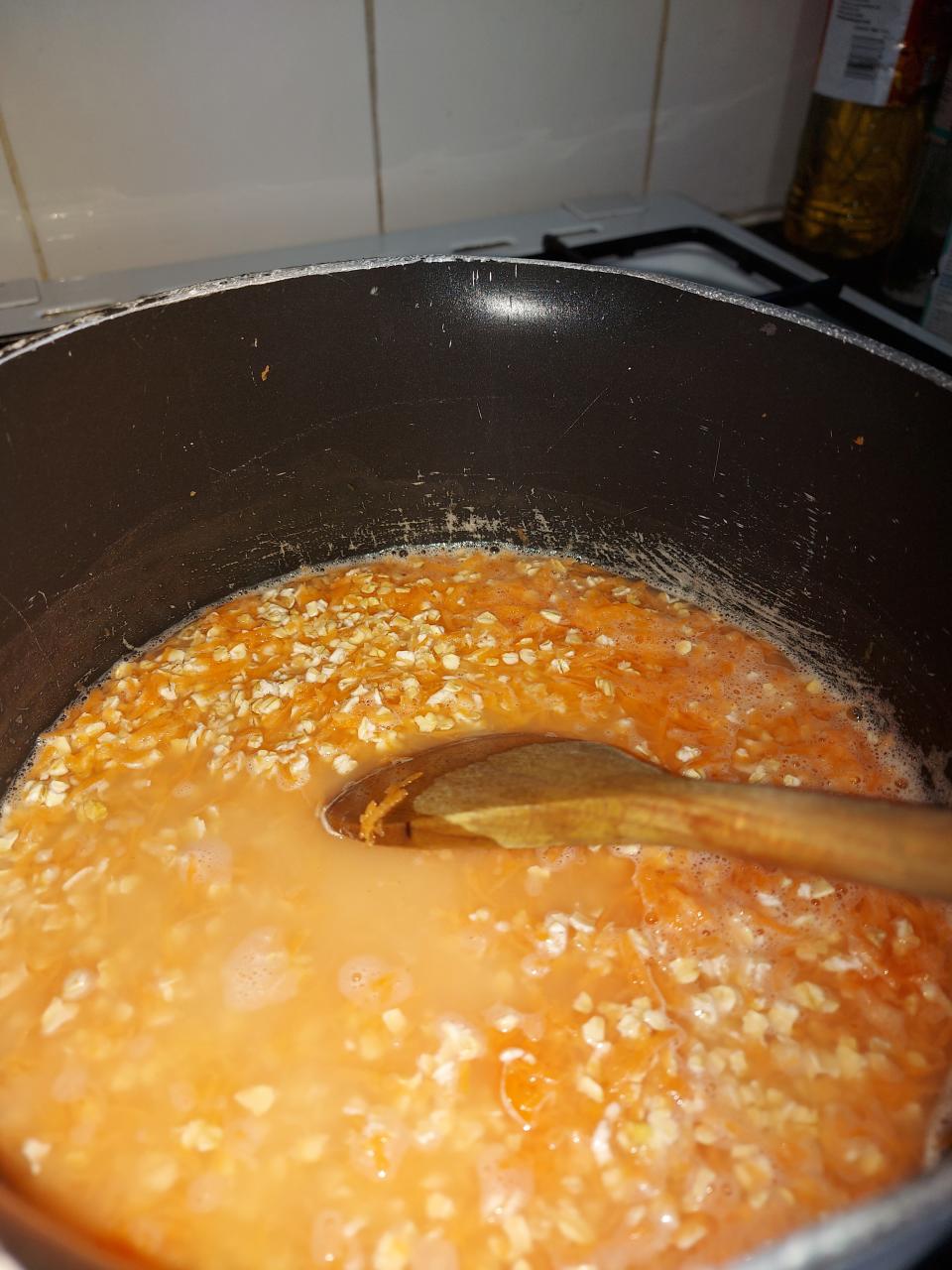 Watery orange oats in a saucepan and a wooden spoon.