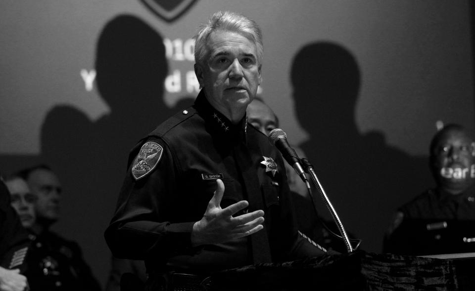 George Gascón, in police uniform, gestures with his hand at a podium with a microphone in front of a screen projection with others seated around him.