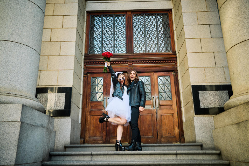 A radiant couple in wedding attire laugh and hold hands against the grand columns of a city building, their joy palpable in the golden sunlight.