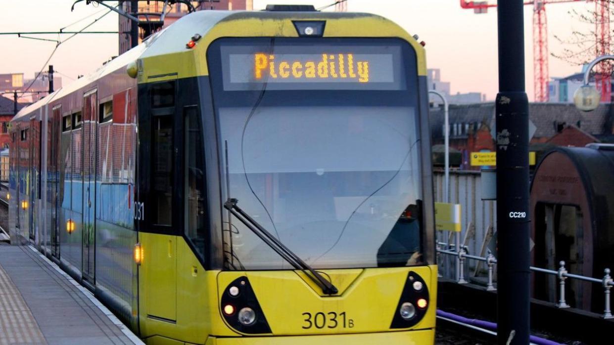 Yellow Metrolink tram at a stop with Piccadilly written on the front