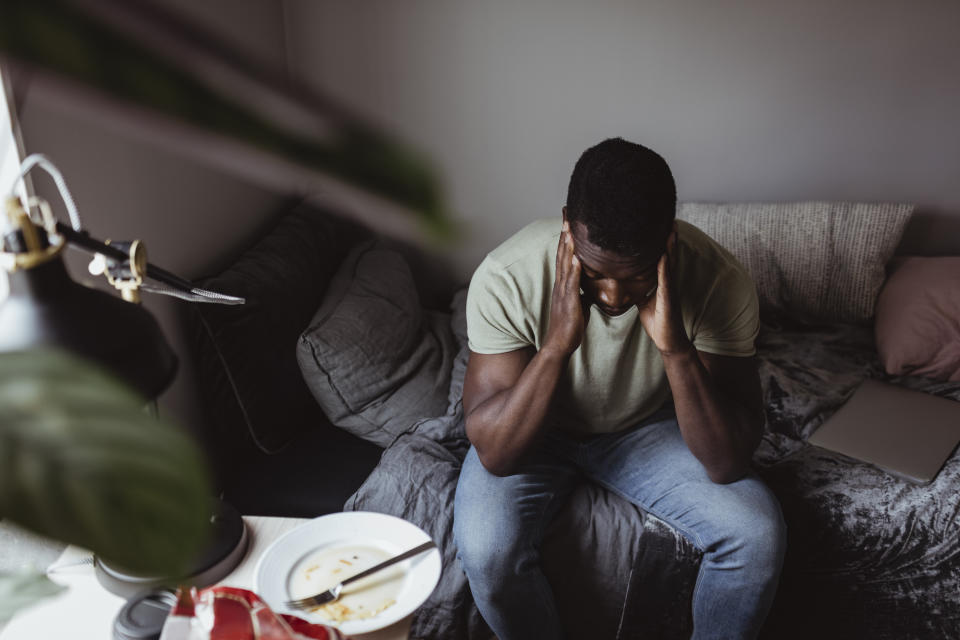 Person sitting on couch with hands on head near an unfinished plate of food, showing signs of distress or headache