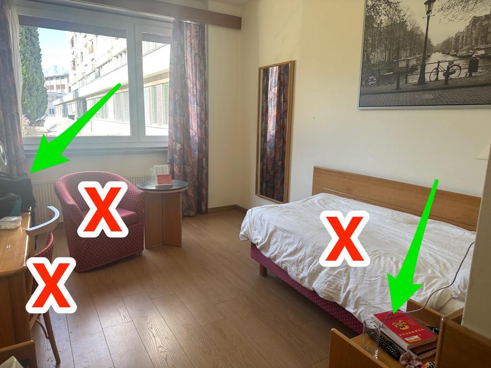 a hotel room with red Xs labeled over the bed, armchair, and upholstered desk chair and green arrows pointing to luggage on the wood desk and bedside table