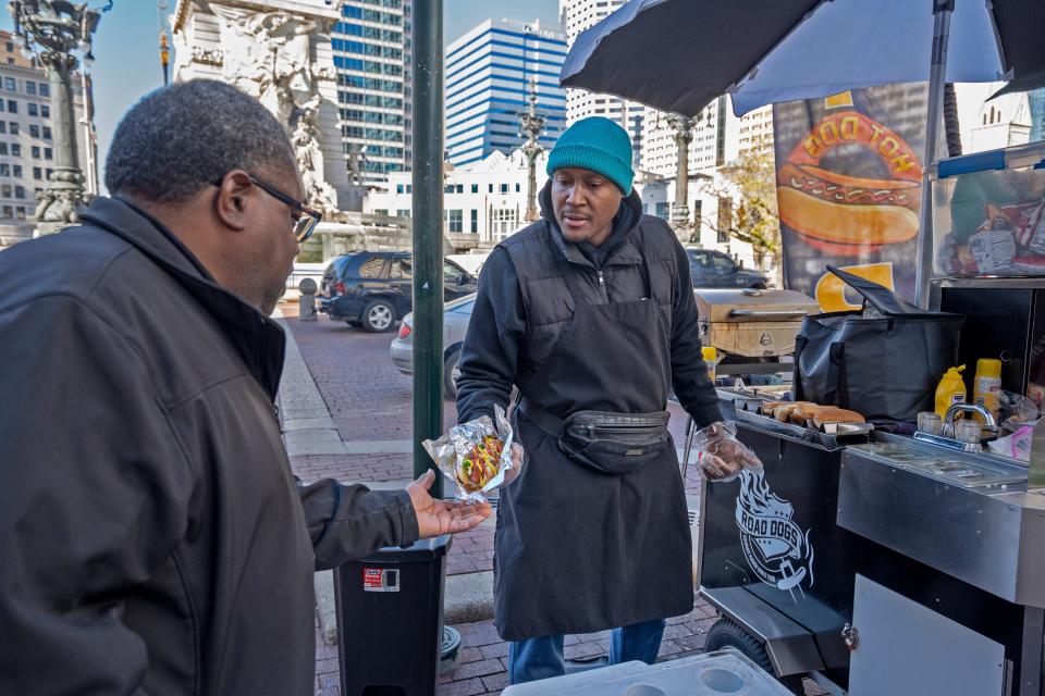 Charles Smith, right, hands Torin Harris a beef polish hot dogs with everyting, from his cart on Monument Circle, Thursday, Nov. 2, 2023. Smith has been in this location since 2019 with his cart, Road Dogs, selling gourmet fusion street food. He loves the freedom and creativitiy of his small business. “A hot dog is a canvas. You can put whatever you want on it to make it special,” he says.