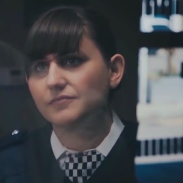 Natasia Demetriou with her hair up in a cop uniform behind a glass booth