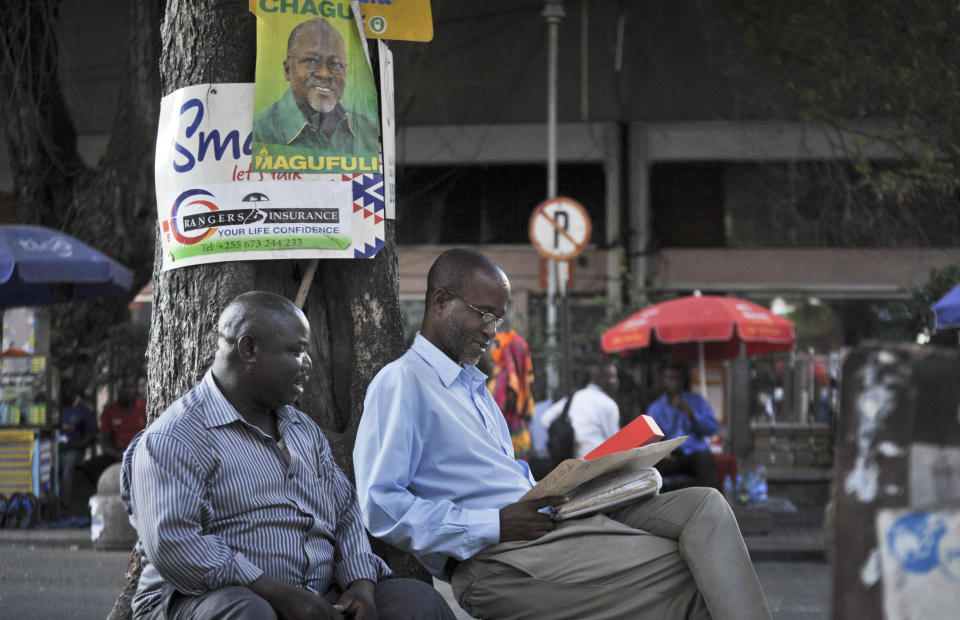 FILE - In this Tuesday, Oct. 27, 2015 file photo, Tanzanians sit next to a tree, underneath an election poster for then ruling party presidential candidate John Magufuli, as they await election results in Dar es Salaam, Tanzania. President John Magufuli of Tanzania, a prominent COVID-19 skeptic whose populist rule often cast his country in a harsh international spotlight, has died aged 61 of heart failure, it was announced Wednesday, March 17, 2021 by Vice President Samia Suluhu. (AP Photo/Khalfan Said, File)