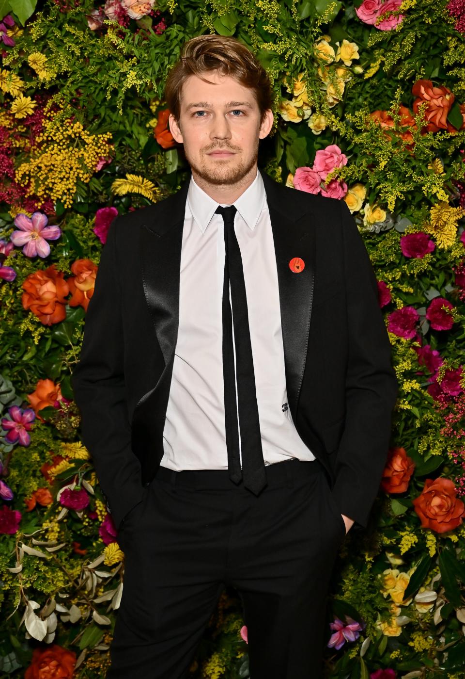 Joe Alwyn standing in front of a floral backdrop wearing a black suit, white shirt, black tie, and red lapel pin
