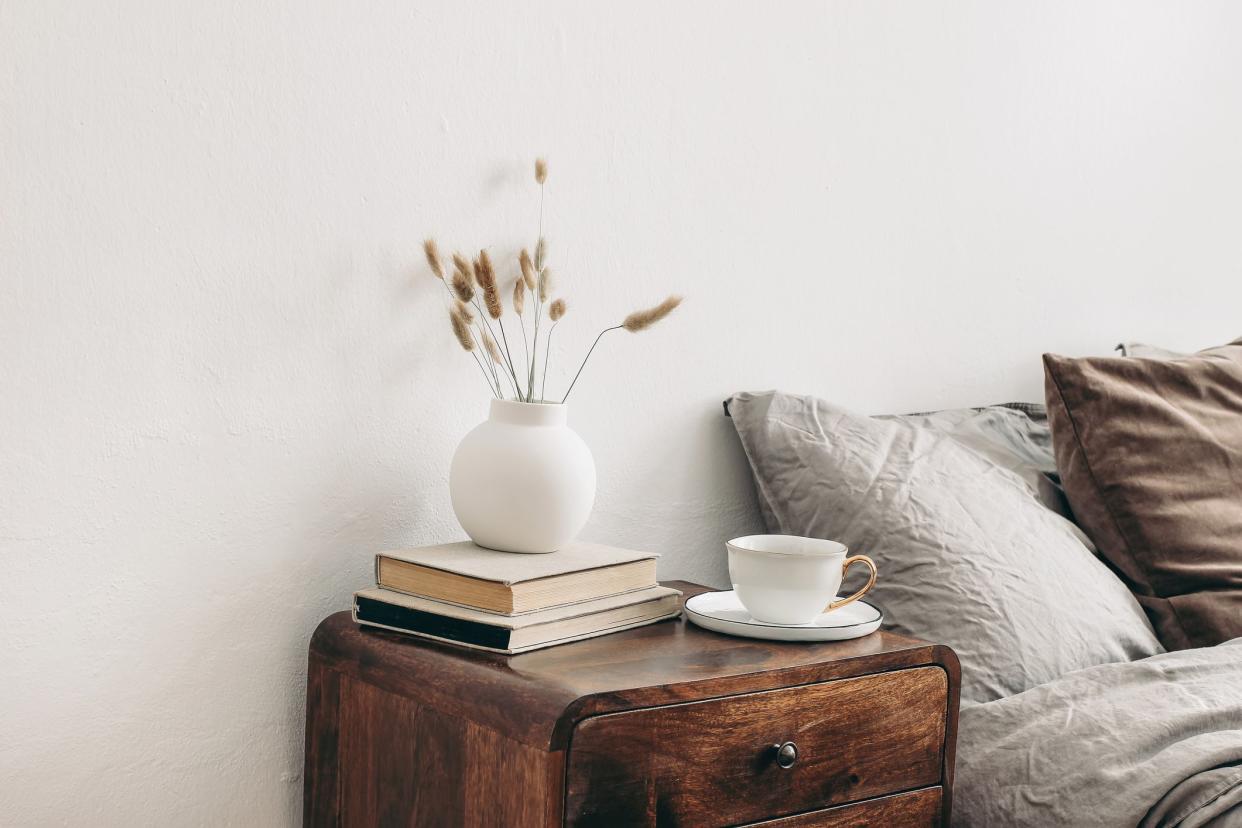 Modern white ceramic vase with dry Lagurus ovatus grass and cup of coffee on retro wooden bedside table. Beige linen and velvet pillows in bedroom, scandinavian interior. Homestaging.