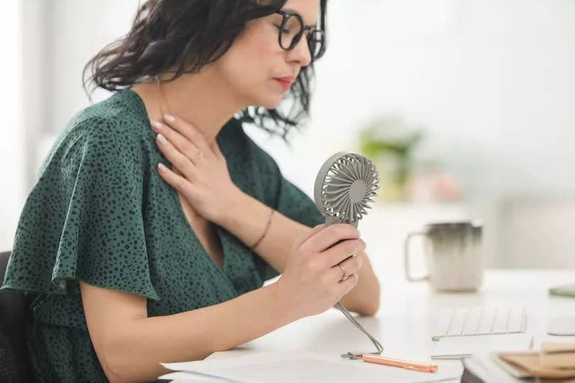 Menopausal mature woman wearing green dress sitting at the desk in the office, having hot flashes and using hand fan.