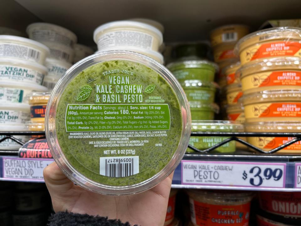 A hand holds a circular container of vegan kale, cashew and basil pesto at Trader Joe's.  The pesto is bright green