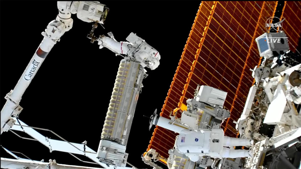 Astronaut Josh Cassada, anchored to the end of the space station's robot arm, manually holds a 750-pound rolled-up solar array blanket while being moved to its installation location. Crewmate Frank Rubio, wearing an unmarked suit, is visible at bottom right. / Credit: NASA TV