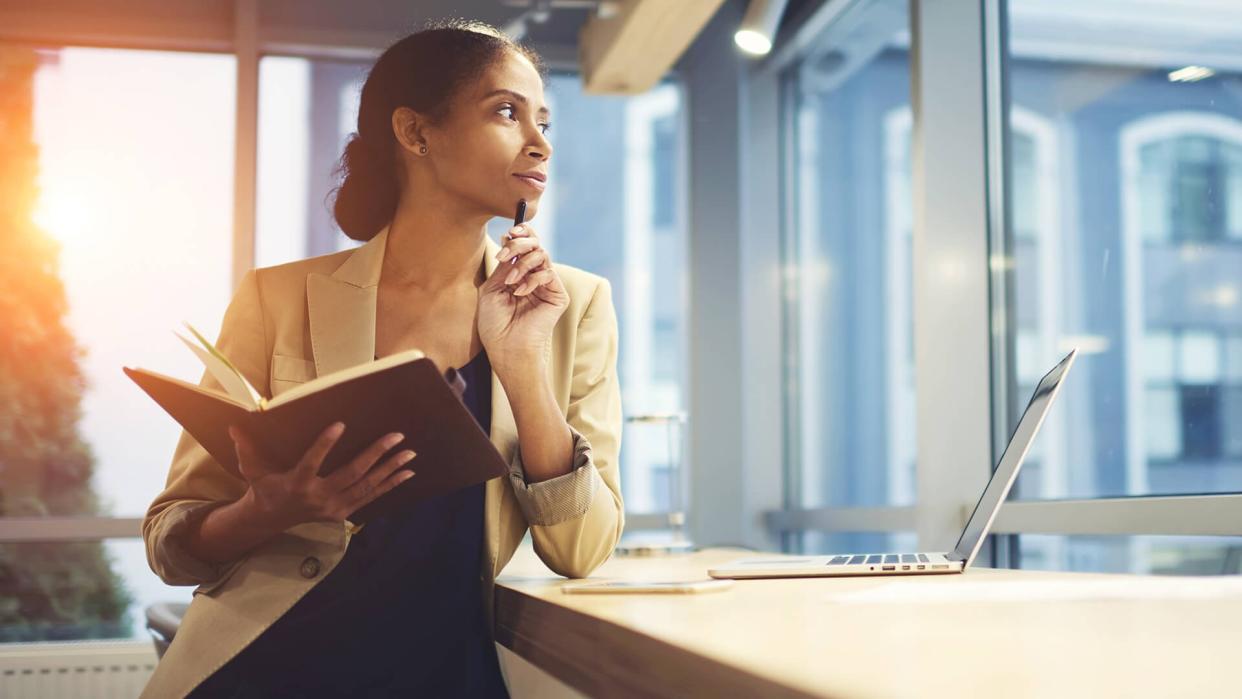 Elegant young woman looking away thoughtfully while holding notepad and leaning on table with laptop.