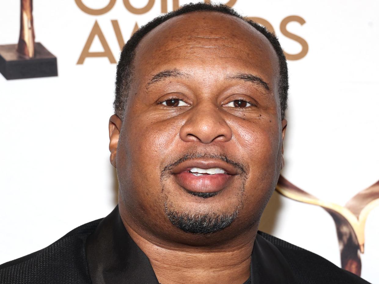 Roy Wood Jr., a man with a trimmed mustache and goatee, smiling on the Writers Guild red carpet. He's wearing a black shirt and a black suit jacket.