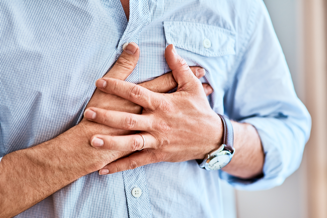 Focus of men's hands gripping his heart area of his chest, wearing a blue shirt, blurred background of white wall on the right