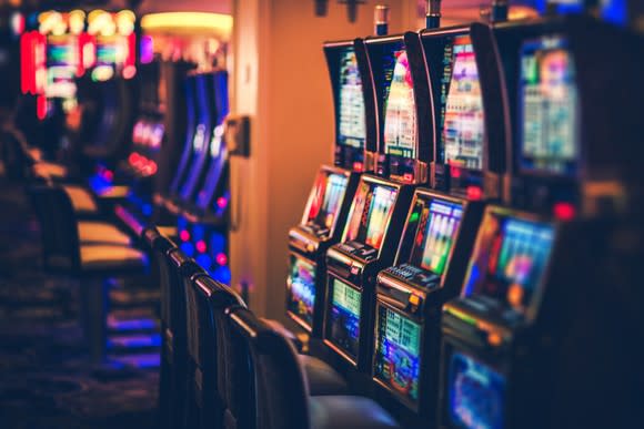 Row of slot machines in a darkened room with Vegas-style lights in the background.