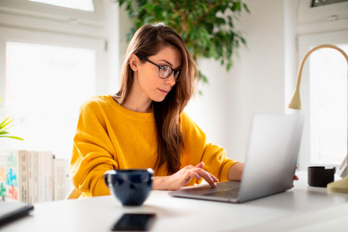 A beautiful young woman enjoying working at home on her laptop in cozy and bright apartment wearing yellow sweater