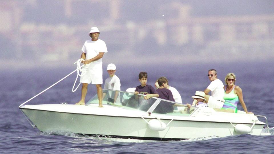 princess diana sits at the back of a boat in a green and blue swimsuit, several other people and children are onboard including a man holding a rope on the front of the boat