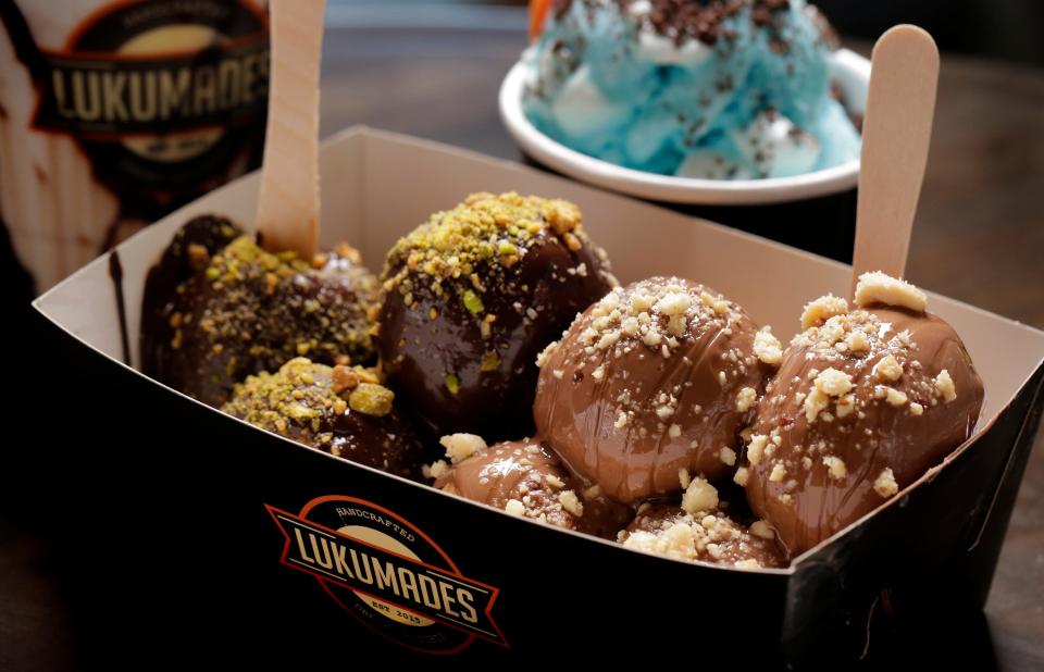 A mixed box of Lukumades Greek doughnuts in flavors Dark Delight, with melted dark chocolate and crushed pistachios, and Snicka Pick, covered with melted milk chocolate, crushed peanuts and drizzled with caramel sauce.
