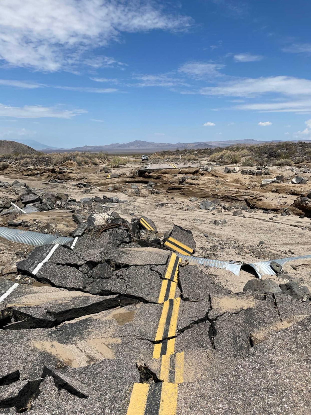 Road asphalt is crumbled up with a metal pipe sticking out.  Clouds loom in the sky over the broken road. Parts of Kelbaker Road have been completely destroyed.