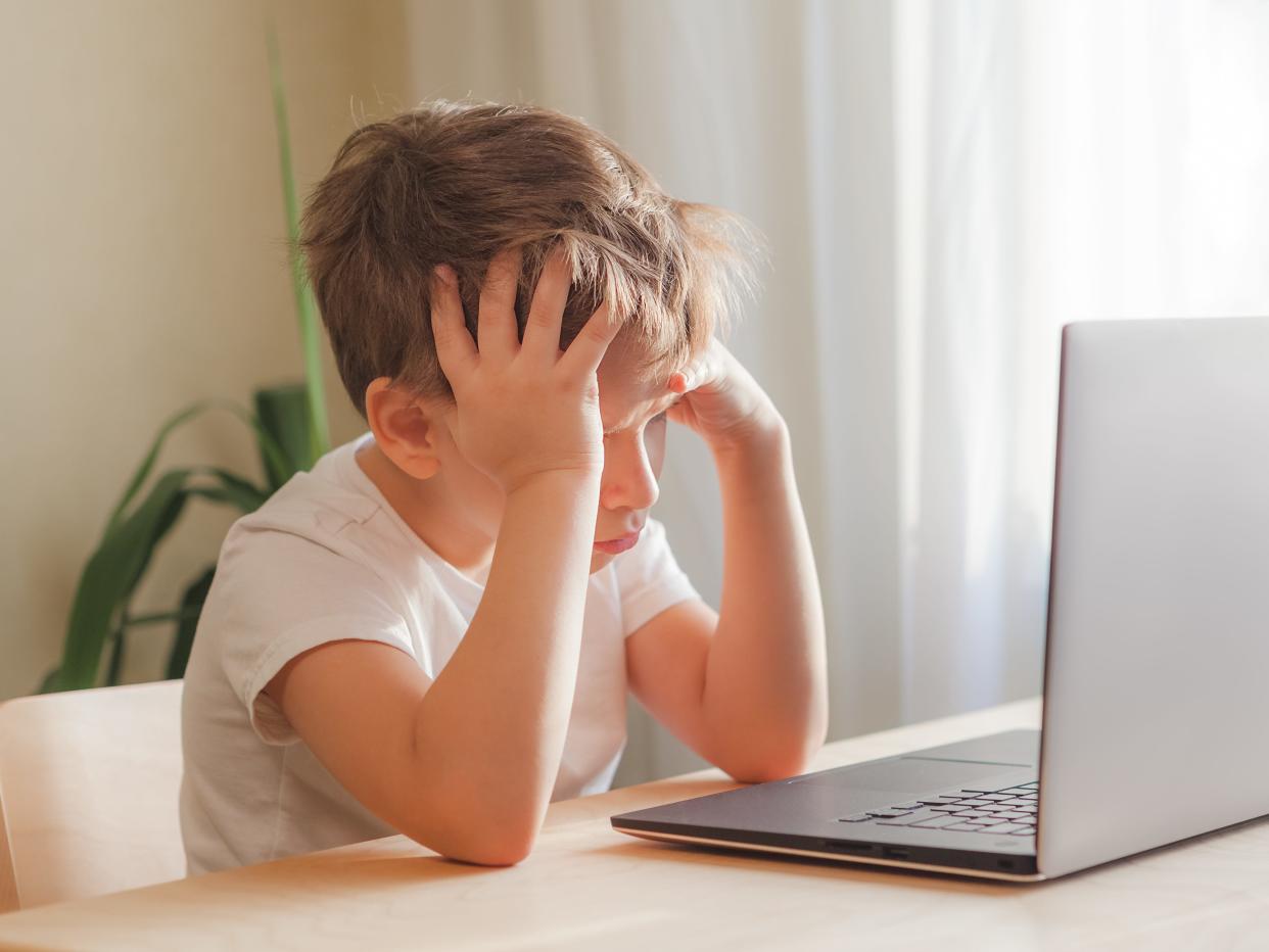 Desperate Child Sits At Desk, Looks Sadly At Computer And Holds His Head With Hands