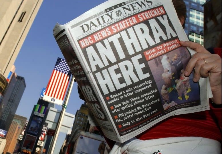 A person in Times Square reads a newspaper with the headline "Anthrax Here." The article mentions NBC News and Tom Brokaw. Yellow taxis and tall buildings are visible