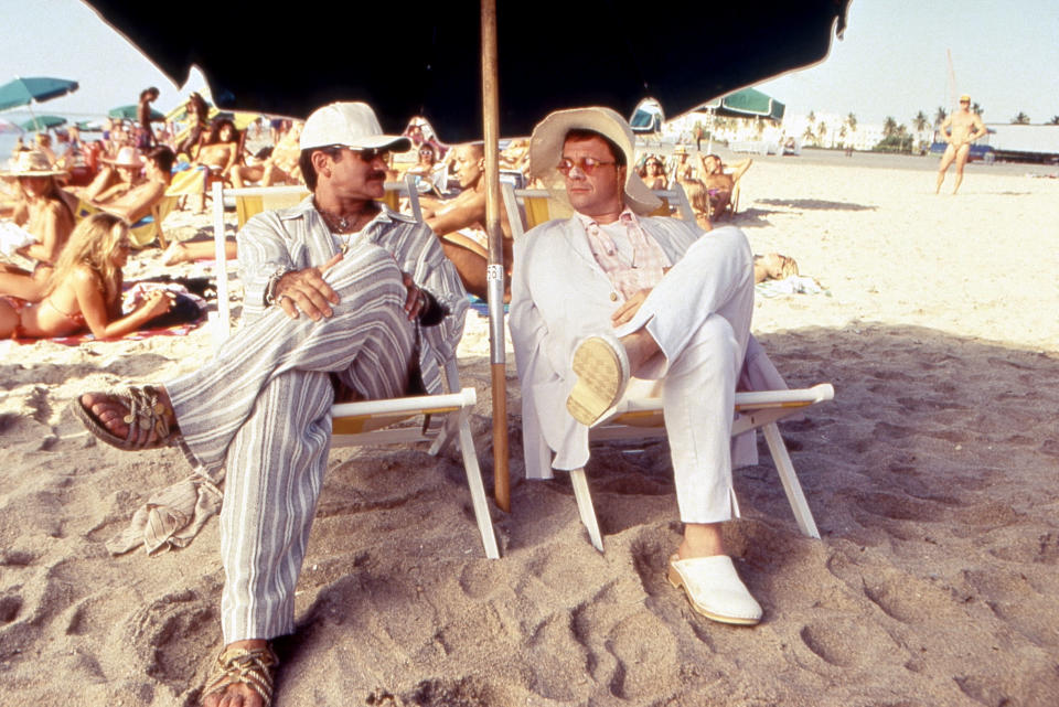 Al Pacino and Robin Williams sit on beach chairs under an umbrella, engaged in conversation, dressed in casual beachwear