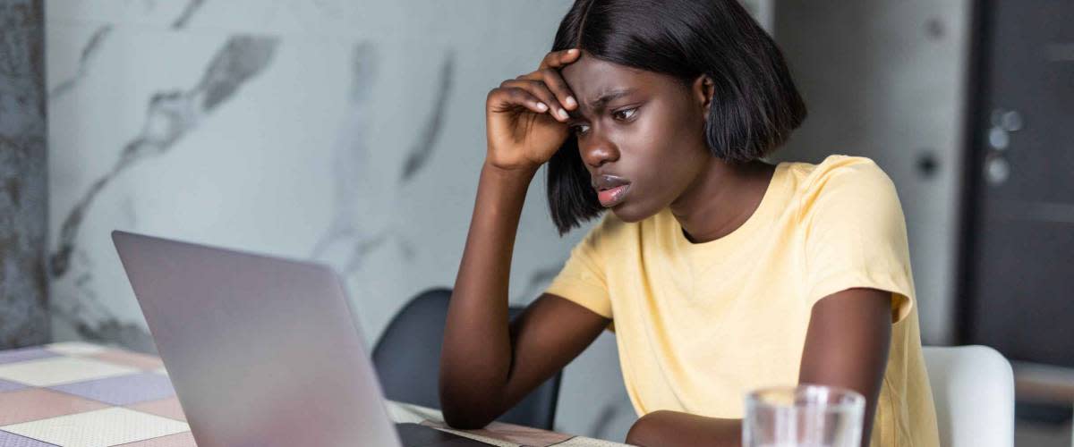 Upset african american female sitting at kitchen table with laptop, dealing with financial stress and pressure because of mortgage debt, worrying a lot or feeling anxious over money
