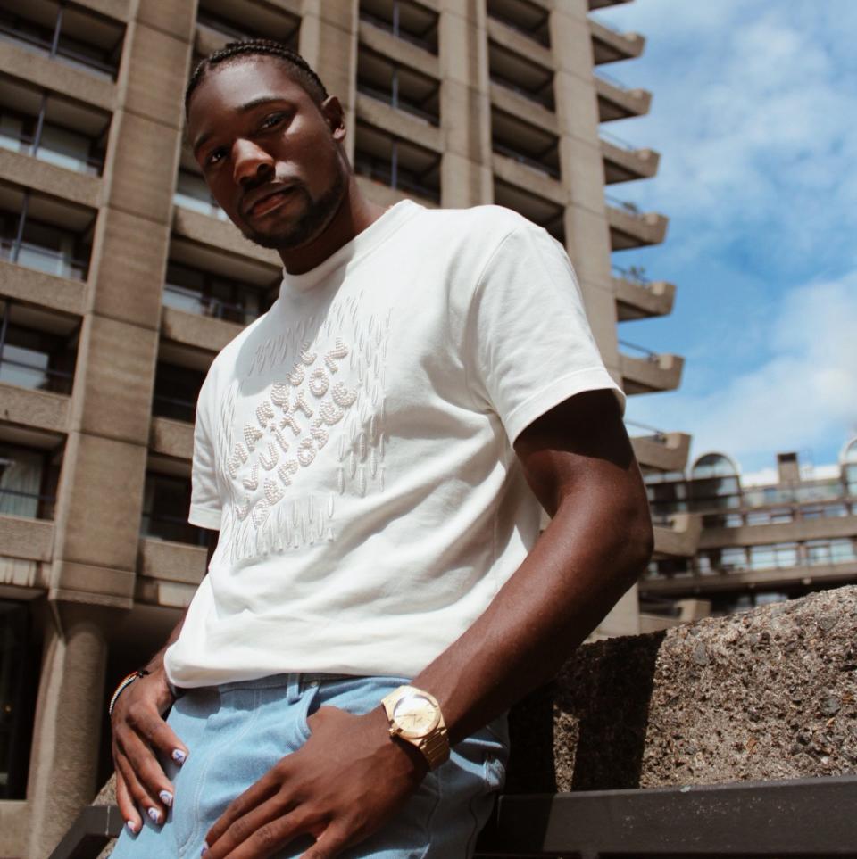 Lyles stood with a tower block in the background wearing a white designer T-shirt and bright blue trousers