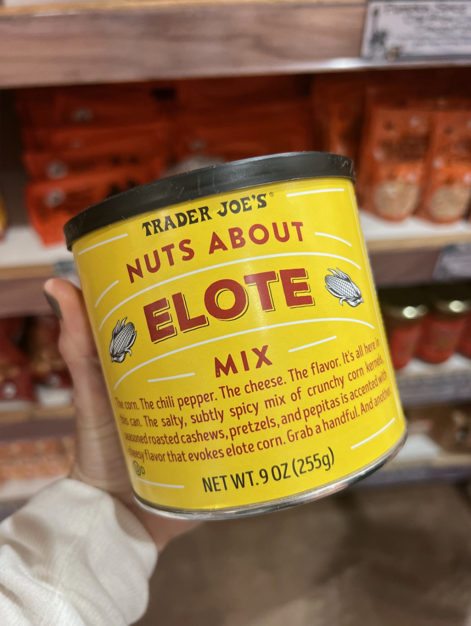 A hand holds a Trader Joe's "Nuts About Elote Mix" can, containing a blend of spicy, cheesy corn kernels, seasoned cashews, pretzels, and pepitas