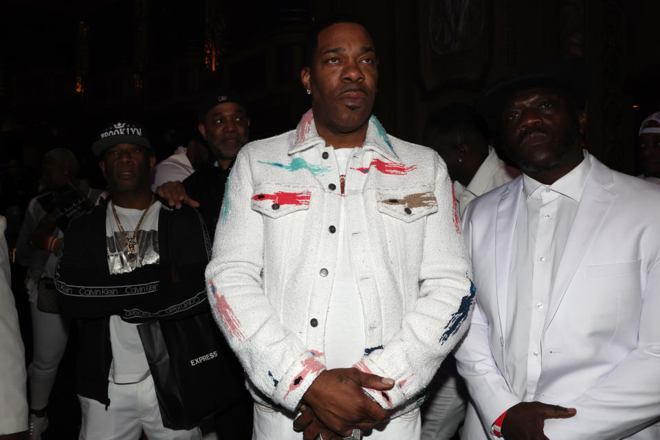 Busta Rhymes, center, at an event wearing a white jacket with colorful accents, surrounded by other individuals in white attire