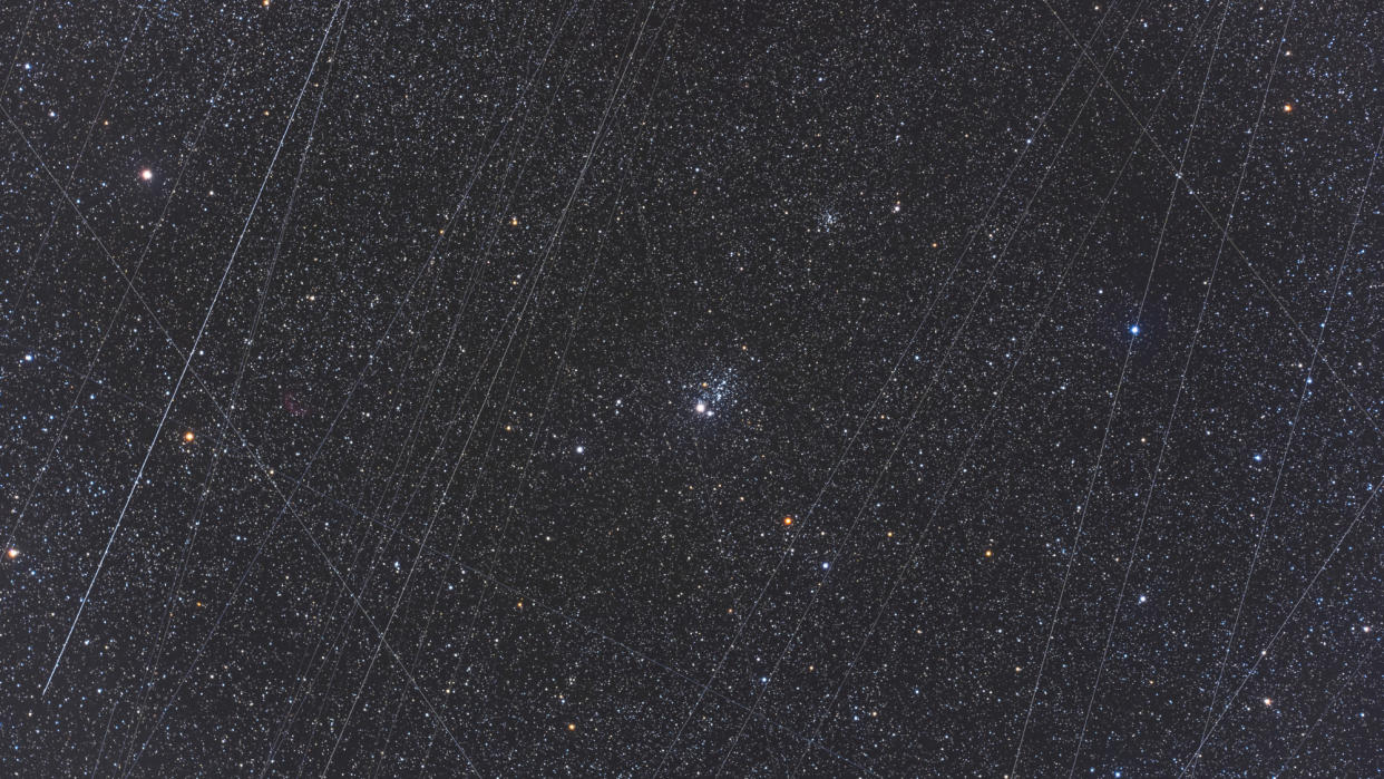  Telescope photo of a star cluster in a dark night sky, with white streaks caused by satellites all over the image. 