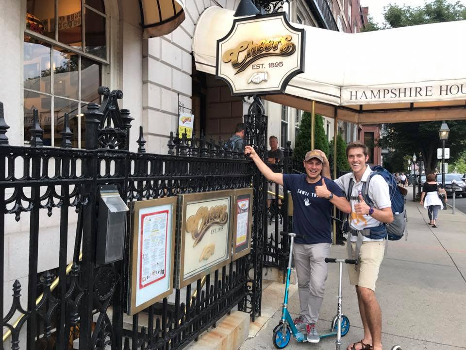 Ash Jurberg and his cousin Yonatan in front of the bar, Cheers, in Boston, and giving the camera the thumbs up. Ash has on a brown baseball hat, navy shirt, grey pants and holds onto the black railing next to him. Yonatan has on a white polo shirt, blue backpack, and tan shorts. They both have scooters parked next to them. 