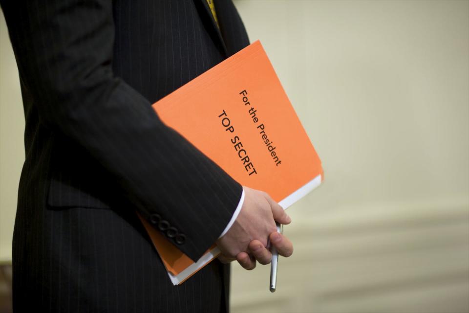 A person, seen from the chest down in a dark suit, holds an orange folder that says 'For the president. Top secret.'