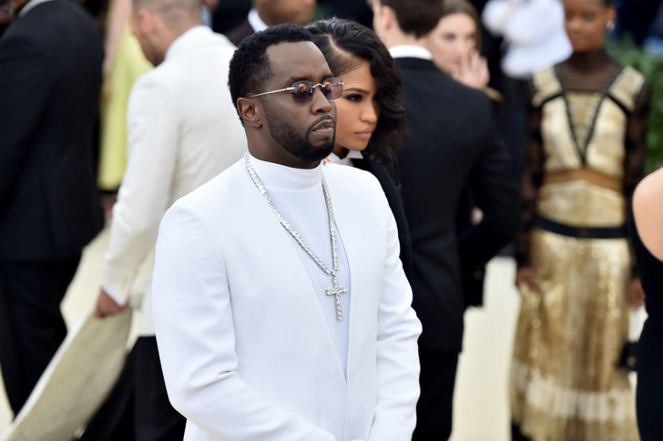 Sean "Diddy" Combs at a red carpet event, wearing a sleek white suit accessorized with a cross necklace