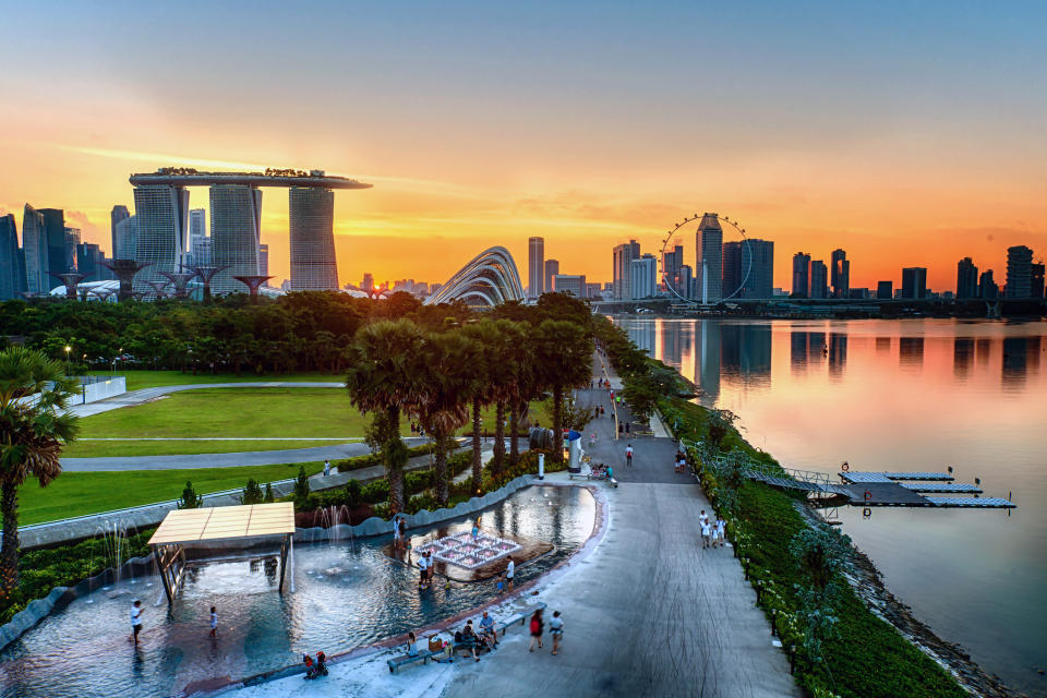 Singapore skyline at sunset with Marina Bay Sands and Singapore Flyer. Gardens by the Bay visible, people walking, and a poolside area in the foreground
