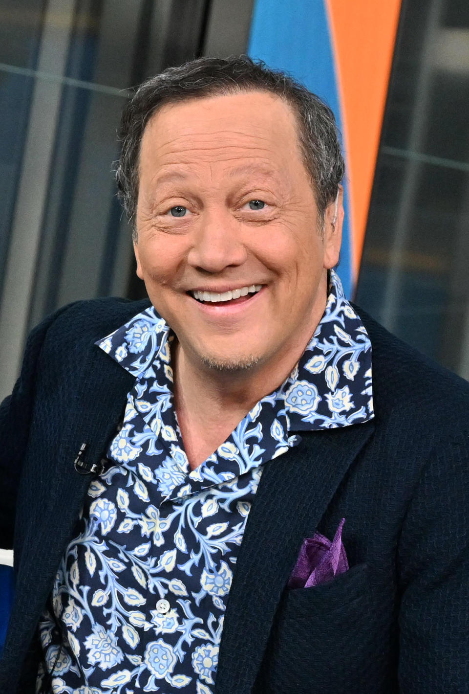 Rob Schneider is seen posing and smiling for the camera in a patterned shirt and black jacket.