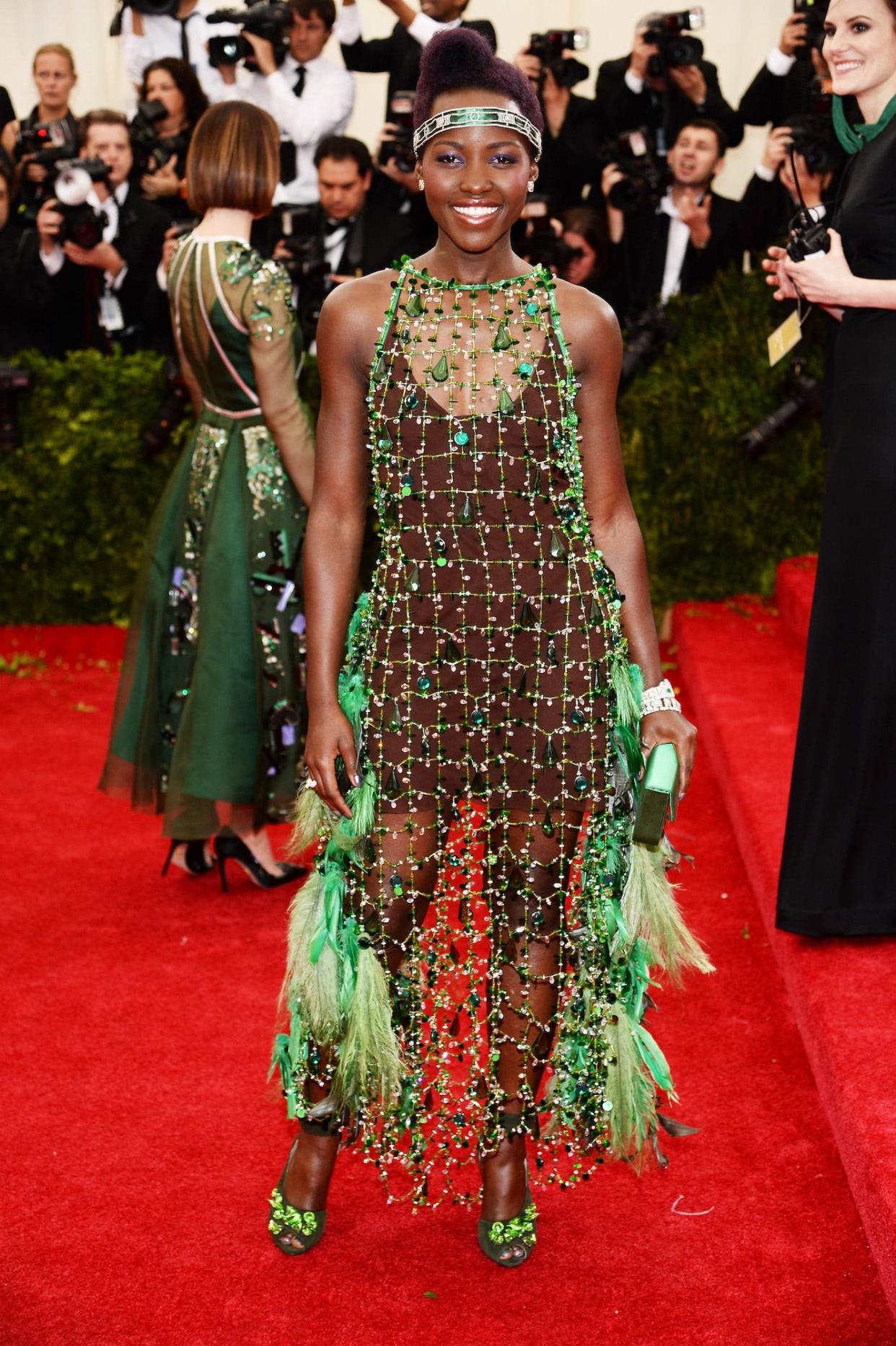Lupita Nyong'o poses on the 2014 Met Gala, wearing a mesh, lime green flapper-style dress decked out with feathers and jewels.