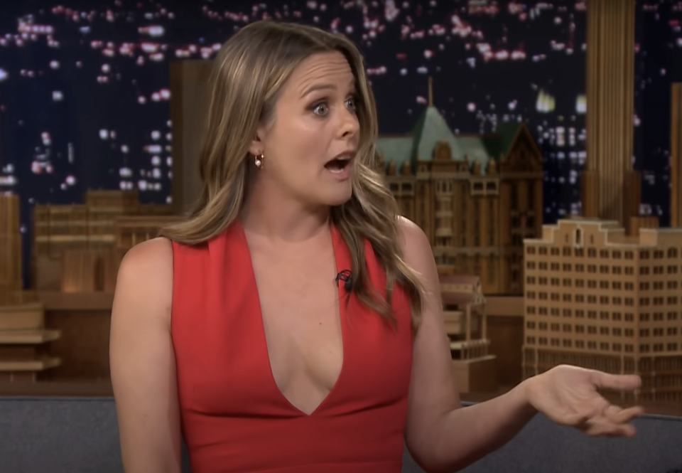 Alicia expressing surprise during a talk show interview, seated with city skyline backdrop