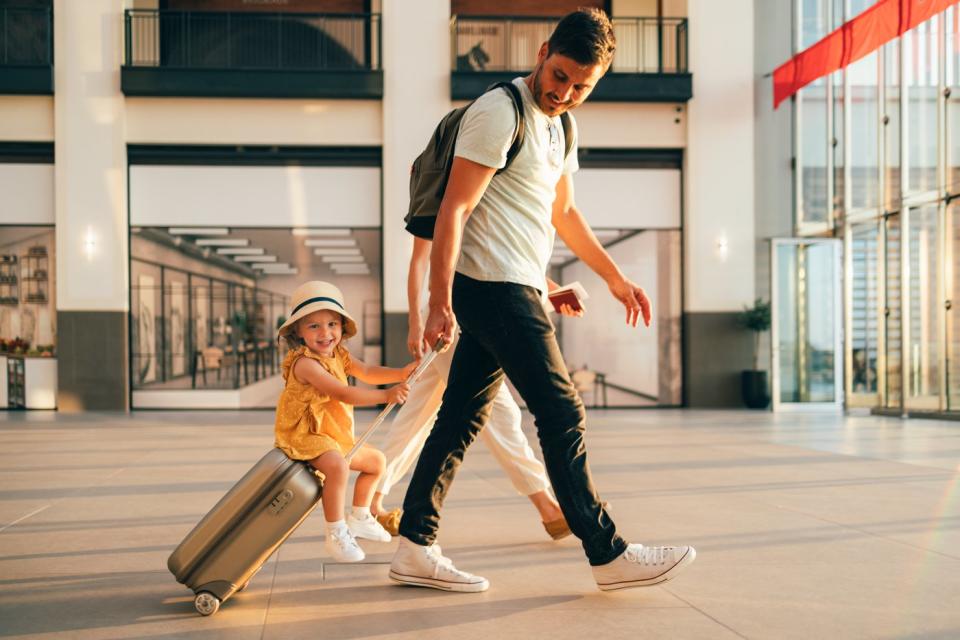 Parent and child walk through an airport, with the child riding on top of a rolling luggage bag.