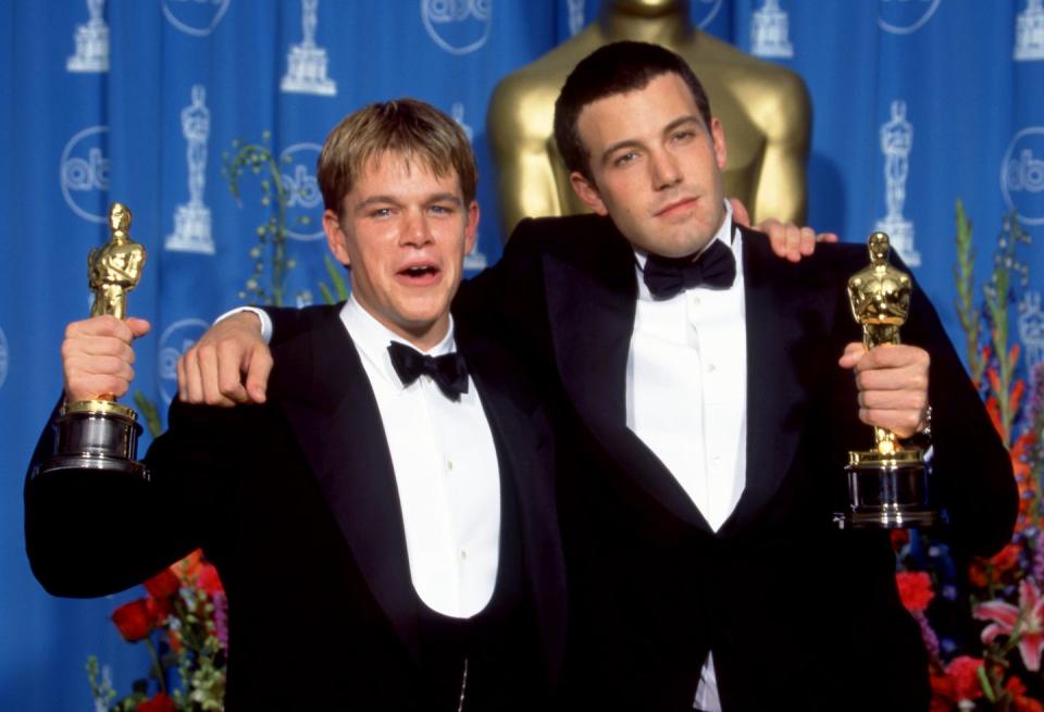 matt damon and ben affleck wearing tuxedos and holding oscar statuettes on a stage, standing in front of a blue curtain and large oscar statue